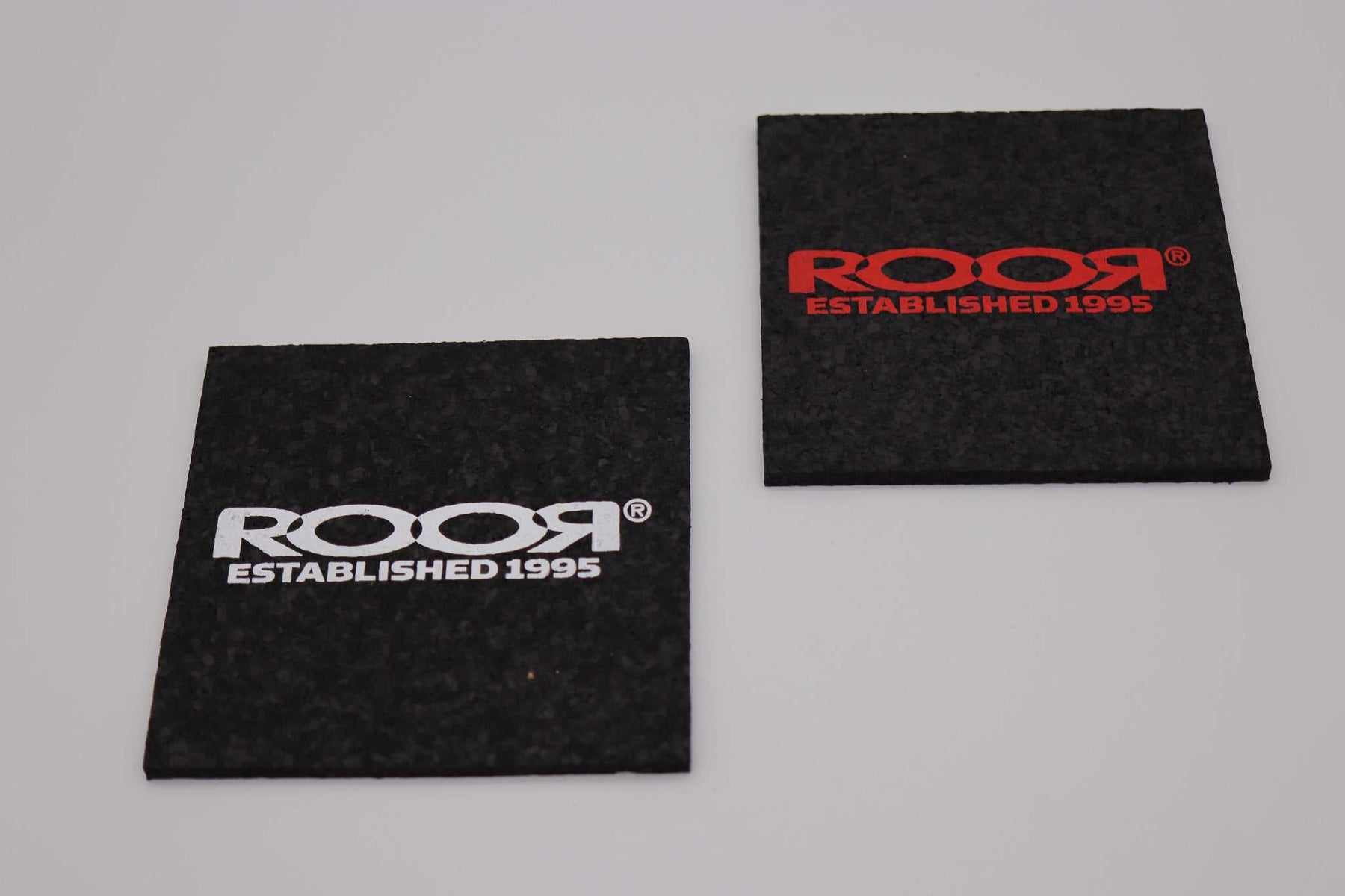 Two black ROOR square coasters with red and white logos protecting surfaces from glass damage