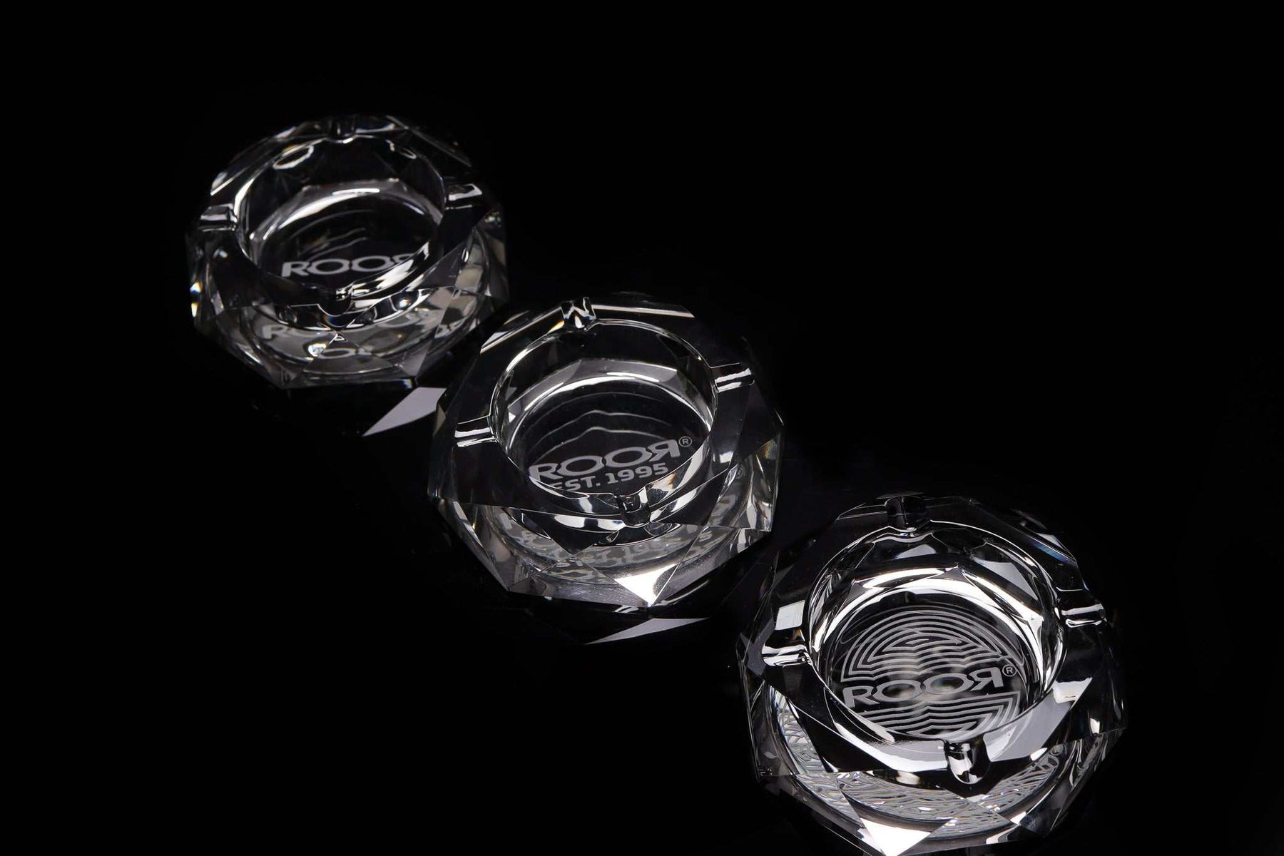 Three clear ROOR glass crystal cut ashtrays with engraved logos on a black background
