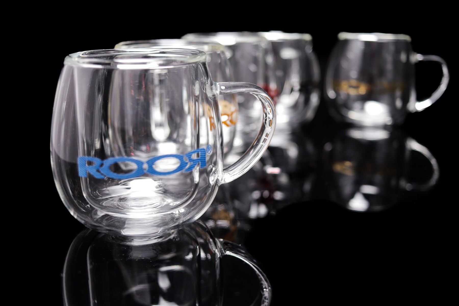 Double-walled ROOR® glass mugs with various decals on reflective surface.